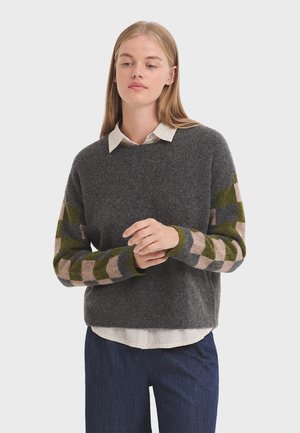 Dunkelgrauer Wollpullover mit ärmeln im Zickzackmuster in Grün und Pink, Rundhalsausschnitt und lockerem Schnitt, getragen über einem weißen Hemd mit Kragen.
