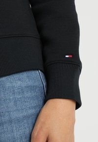 Czarna bluza z prążkowanymi mankietami, wykonana z miękkiego materiału. Charakterystycznym elementem jest małe logo w kolorach czerwonym, białym i niebieskim na rękawie.