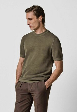 Maglione a maniche corte verde oliva a costine con scollo rotondo, abbinato a pantaloni marroni. Tessuto lavorato a trama e polsini a contrasto.