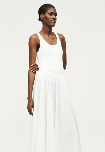 Νεαρή γυναίκα με σκούρο δέρμα και κοντά μαλλιά, φοράει λευκή αμάνικη μπλούζα και μακριά λευκή φούστα, στέκεται με το ένα χέρι σταυρωμένο.