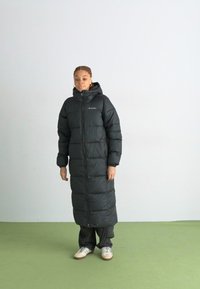 Černý dlouhý zateplený kabát s vysokým límcem a předním zipem, navržený pro teplo. Model stojí na světlém pozadí se zelenou podlahou.