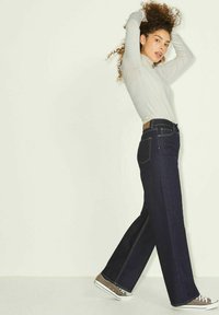 Jeans de mezclilla oscura de pierna ancha con detalles en las costuras laterales, llevados con una camiseta de manga larga gris claro y zapatillas marrones.