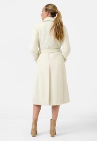 Long cream coat with a belt, featuring a notched collar and back vent. Worn with beige heeled boots, showcasing a smooth texture.
