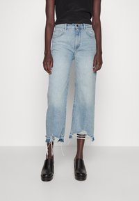 Jeans large bleu clair, à ourlets effilochés, avec un design à cinq poches et une fermeture à bouton en laiton, associés à des sabots à plateforme noirs.