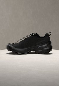 Μαύρο παπούτσι trail running Salomon με κορδόνι έλξης, ανθεκτική σόλα και σύστημα γρήγορου δέσιμου, σε ελαφριά στρογγυλή πλατφόρμα με γκρι φόντο.
