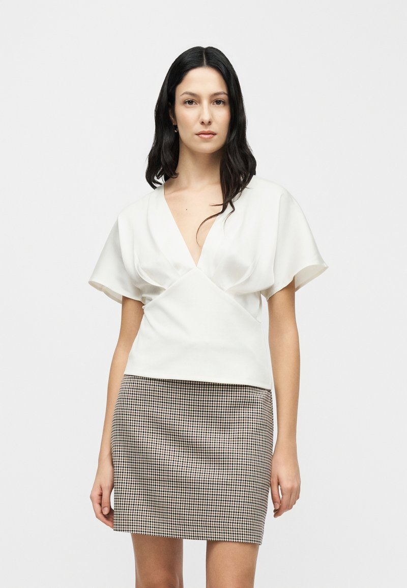 Haut blanc à manches courtes avec un décolleté en V profond, design portefeuille, associé à une mini-jupe en pied-de-poule marron et noir. Texture lisse.