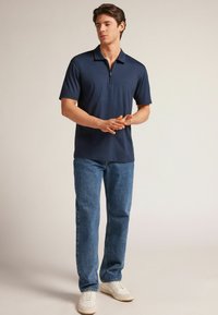 Granatowa koszulka polo z krótkimi rękawami i zamkiem w kołnierzu, w zestawieniu z jasnoniebieskimi prostymi dżinsami i białymi trampkami. Gładka tekstura materiału.