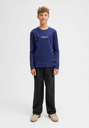 Námornícka košeľa s dlhým rukávom a logom, čierne nohavice a béžové slip-on topánky, ktoré nosí dieťa s kučeravými vlasmi. Jednoduchý, neformálny dizajn.