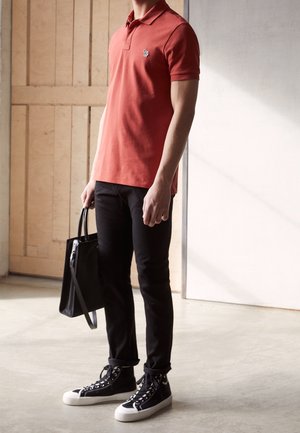 Person trägt rostfarbenes Polo, schwarze enge Hose und schwarze High-Top-Sneaker, hält eine schwarze Tragetasche in einem lichtdurchfluteten Raum.