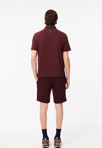 Bordeaux polo shirt met een kraag, gecombineerd met bijpassende shorts. Het shirt heeft een groene tag aan de achterkant. Het materiaal lijkt soepel en casual.