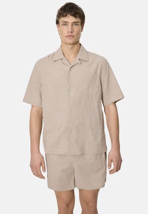 Hombre vestido con camisa beige de manga corta con botones y pantalones cortos a juego, de pie mirando hacia adelante contra un fondo liso.