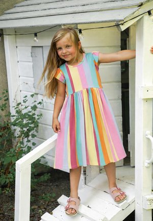 Junges Mädchen in einem bunten gestreiften Kleid steht auf weißen Holzstufen, hält das Geländer, lächelt, mit grünen Pflanzen in der Nähe.