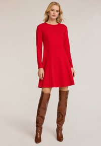 Rotes langärmliges Kleid mit tailliertem Oberteil und fließendem Rock, kombiniert mit braunen kniehohen Stiefeln, die eine glatte Textur und ein schlichtes Design zeigen.