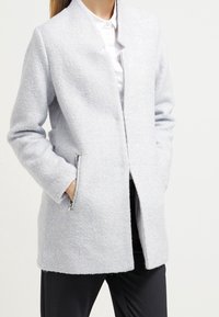 Cappotto grigio chiaro, sartoriale, realizzato in tessuto testurizzato con colletto ad ascot, tasche laterali con zip e taglio dritto. Adatto per un abbigliamento casual.