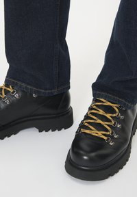 Stivali in pelle nera con suole spesse e lacci gialli, indossati con jeans in denim blu scuro su uno sfondo bianco.