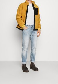 Giacca di pile gialla con dettagli neri, t-shirt grigia, jeans azzurri chiari e stivali alla caviglia marroni. La giacca presenta una chiusura con zip e tasche.
