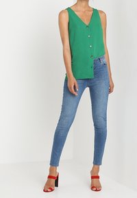 Grön ärmlös blus med V-ringning och knapprad, tillsammans med blå smala jeans och röda klacksandaler med blockheel.