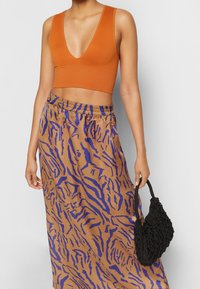 Hnědý crop top s hlubokým V-výstřihem; hnědá maxi sukně s fialovým proužkovým vzorem tygra; nese černou pletenou kabelku.