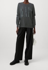 Felpa grigia con il logo "GAP" in lettering lucido, abbinata a pantaloni a gamba larga neri a strisce e stivali alla caviglia neri, mentre tiene una borsa bianca.