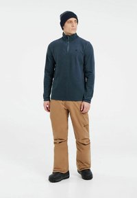 Jūros jėgos vilnos puloveris su pusiau užtrauktuku ir aukštu apyvaišu, derinamas su rudomis vandeniui nepralaidžiomis kelnėmis ir juodais batais. Matoma tekstūruota audinio detalė.