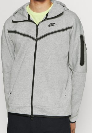 Mężczyzna ubrany w jasnoszarą bluzę Nike zapinaną na zamek z czarnymi akcentami i kieszenią na zamek na lewym rękawie, w połączeniu z żółtą koszulką pod spodem.