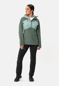 Giacca leggera con cappuccio di colore verde scuro e teal, dotata di tasche con zip. Abbinata a pantaloni neri con sezioni testurizzate sulle ginocchia e calzature scure.