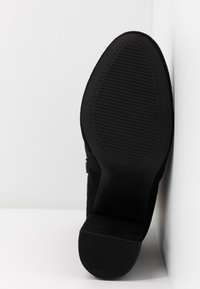 Semelle de chaussure en daim noir avec une semelle texturée antidérapante et un petit talon carré, positionnée debout contre un mur d'angle blanc.