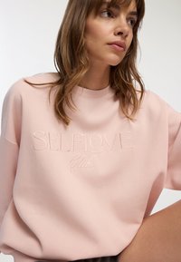 Jasnoróżowy sweatshirt wykonany z miękkiej tkaniny, z wyszywanym wzorem "SELFLOVE Club". Krój jest luźny i ma okrągły dekolt.