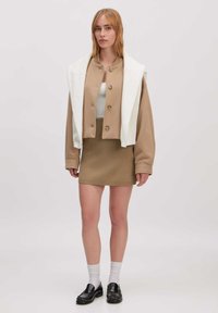 Afgeknot beige jasje met knopen en een witte oversized kraag, gelaagd over een aansluitende beige rok. Zwarte loafers en grijze sokken maken de look compleet.