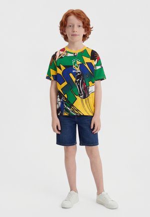 Αγόρι με κόκκινα μαλλιά που φοράει πολύχρωμη μπλούζα με γραφικό στοιχείο Βραζιλίας, σκούρο τζιν σορτς και λευκά αθλητικά παπούτσια, στέκεται μπροστά σε απλό φόντο.