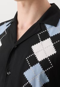 Homme portant une chemise noire avec un motif argyle en zigzag bleu et blanc, ornée de boutons noirs, montrant le col et le haut de la poitrine.