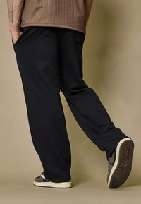 Personne portant un pantalon noir ample, un haut côtelé marron et des baskets noir et blanc, se tenant avec un pied levé contre un fond beige uni.