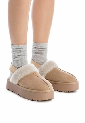 Pantofole in suede beige con un morbido bordo in pelliccia sintetica bianca, dotate di una spessa suola piatta beige e calze grigie a coste.