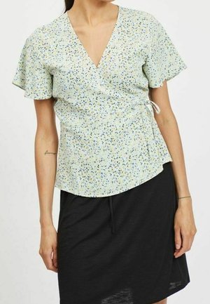 Femme portant un chemisier cache-cœur à fleurs vert clair avec des manches courtes volantées et une jupe noire longueur genou.