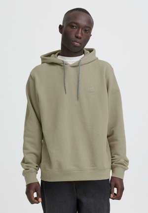 Sweat à capuche vert olive en tissu texturé, avec capuche à cordon de serrage et petit logo embossé sur la poitrine. Associé à un pantalon gris foncé.