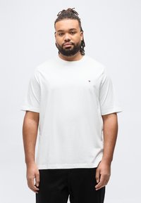 Άνδρας με κόμπους μαλλιών (dreadlocks) και γενειάδα, φορώντας ένα απλό λευκό μπλουζάκι με μικρό λογότυπο στο στήθος, στέκεται μπροστά σε λευκό φόντο.