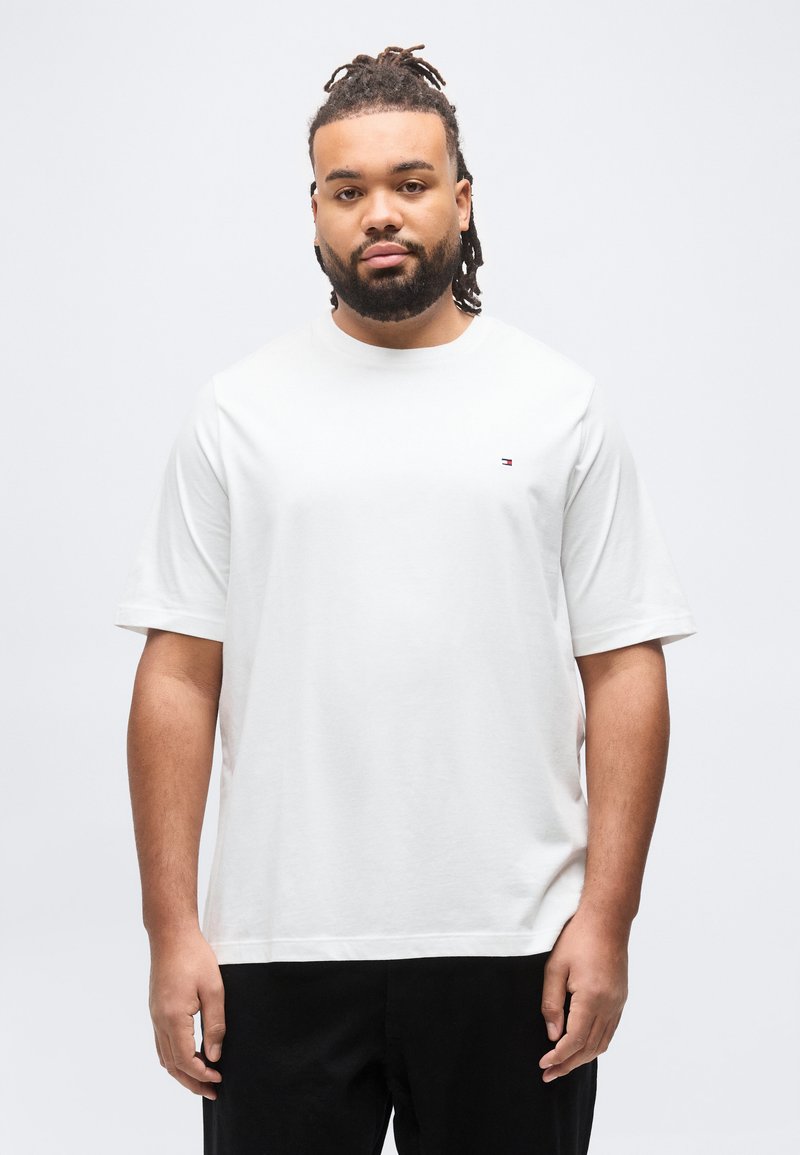 Άνδρας με κόμπους μαλλιών (dreadlocks) και γενειάδα, φορώντας ένα απλό λευκό μπλουζάκι με μικρό λογότυπο στο στήθος, στέκεται μπροστά σε λευκό φόντο.