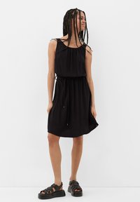 Schwarzes ärmelloses Kleid mit gerafftem Ausschnitt, verstellbarem Kordelzug in der Taille und schwingendem Saum. Kombiniert mit klobigen schwarzen Sandalen.