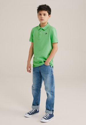 Camisa polo verde de mangas cortas, con tapeta de tres botones y pequeño logo negro; combinada con unos vaqueros claros remangados y zapatillas en color azul marino.