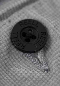 Black round button with "REELL" embossed, attached to textured gray fabric with small stitches in a V-shape near the button.