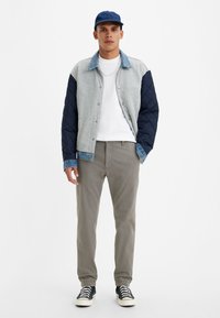 Giacca trapuntata grigia e blu navy, colletto in denim; t-shirt bianca sotto; pantaloni khaki, scarpe nere con suole bianche, cappellino blu.