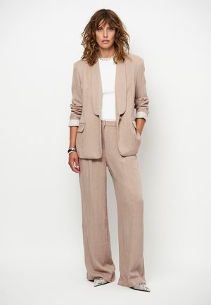 Vrouw met golvend haar draagt een beige blazer en wijde broek, witte top, zilveren ketting en armband, en puntige schoenen met slangenprint, staand.