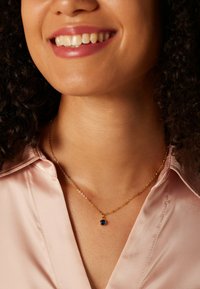 Gouden ketting met een kleine blauwe hanger, gedragen over een lichtroze satijnen blouse, die de soepele textuur en kleurcontrasten benadrukt.