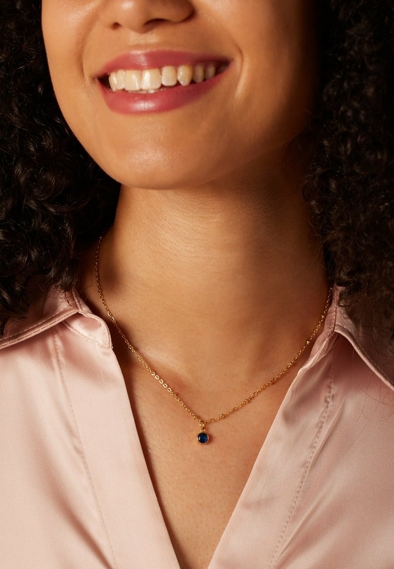 Gouden ketting met een kleine blauwe hanger, gedragen over een lichtroze satijnen blouse, die de soepele textuur en kleurcontrasten benadrukt.