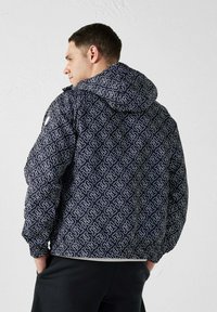 Uomo con giacca nera con cappuccio con motivo di lettere bianche intrecciate, in piedi con le mani in tasca, di spalle e leggermente inclinato.