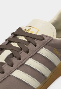 Κοντινό πλάνο σε καφέ σουέτ αθλητικό παπούτσι με κρεμ δερμάτινες ρίγες, κορδόνια στο ίδιο χρώμα και χρυσό λογότυπο στη γλώσσα και στο πλάι.