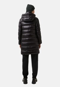 Osoba w czarnej pikowanej kurtce z kapturem, czarnych spodniach cargo, czarnych butach i ciemnozielonej dzianinowej czapce, odwrócona tyłem na białym tle.