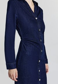 Robe chemise en denim navy à manches longues, devant boutonné et détail froncé sur le côté. Comprend un col classique et des boutons à ton argent.