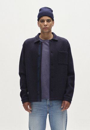 Sins tinu jaka, kas izgatavota no aviācijas vilnas maisījuma, ar pogu aizdari, krūšu kabatu un kontrastējošu rildu. Uzvilkta virs gaiši purpursarkanām krekla un džinsa.