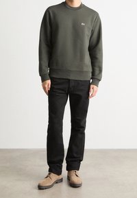 Olive green sweatshirt with a crew neck and embroidered logo, paired with black jeans and tan suede shoes, standing on a neutral floor.